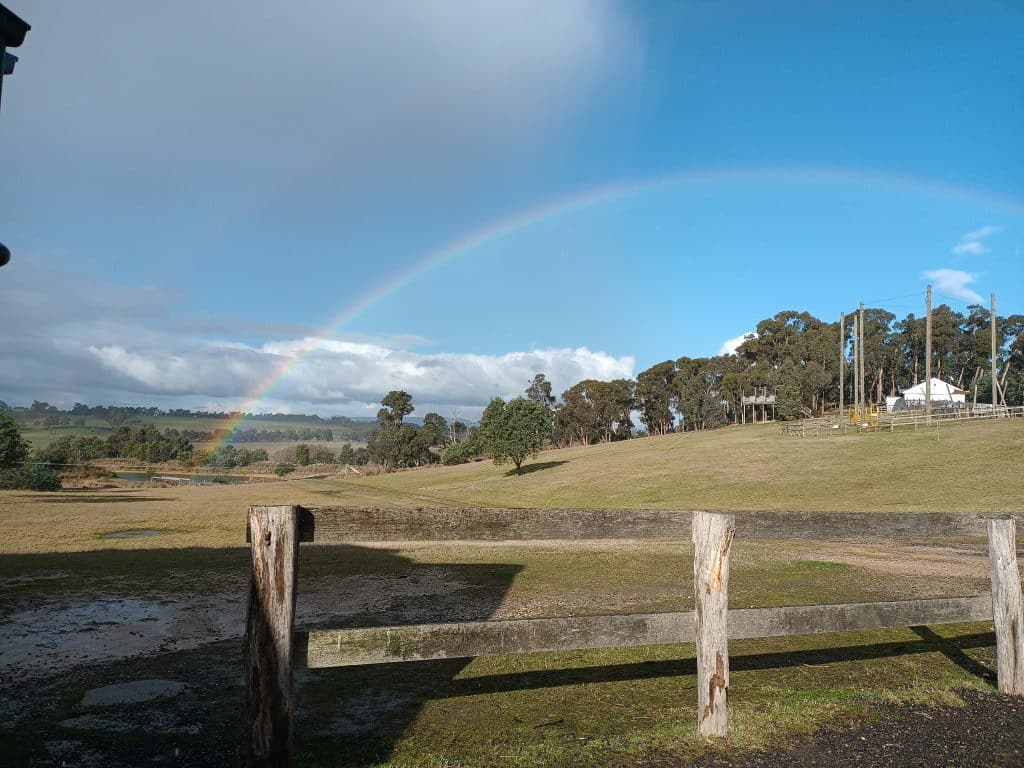 A rainbow arcs over a grassy field with scattered trees, viewed from behind a wooden fence under a partly cloudy sky at a PGL camp.