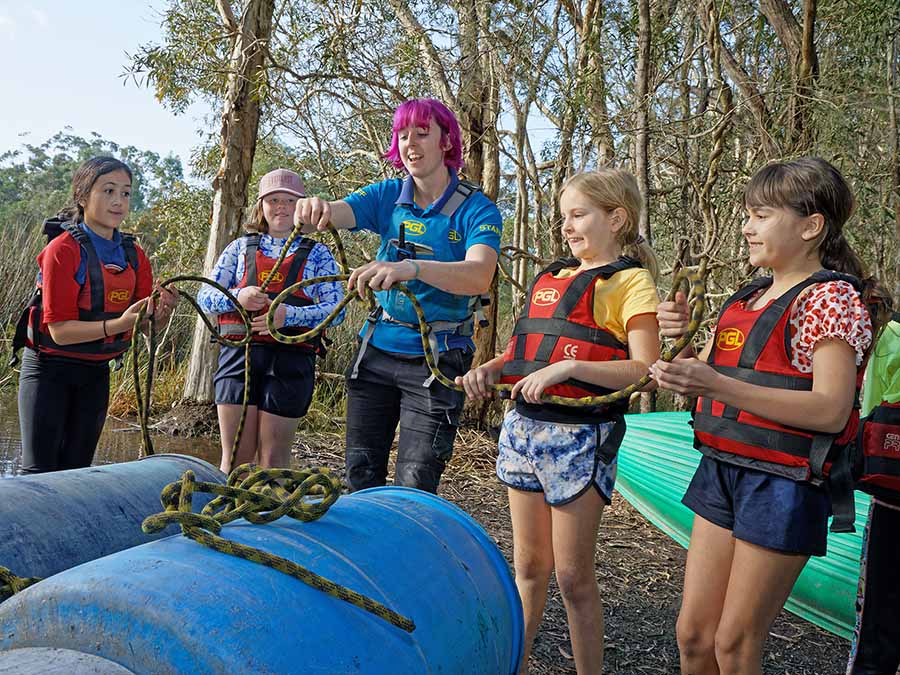 A group of children wearing life jackets learn to tie knots on a kayak under the guidance of an instructor with pink hair.