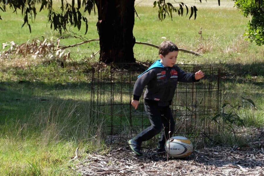 A young boy in a wetsuit playing with a rugby ball near a tree and wire fencing in a grassy field.