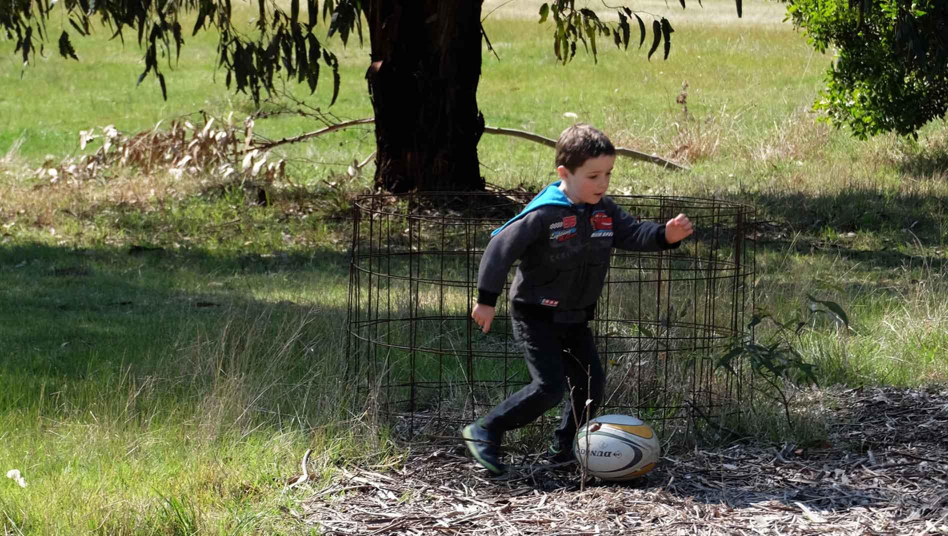 A young boy in a wetsuit playing with a rugby ball near a tree and wire fencing in a grassy field.