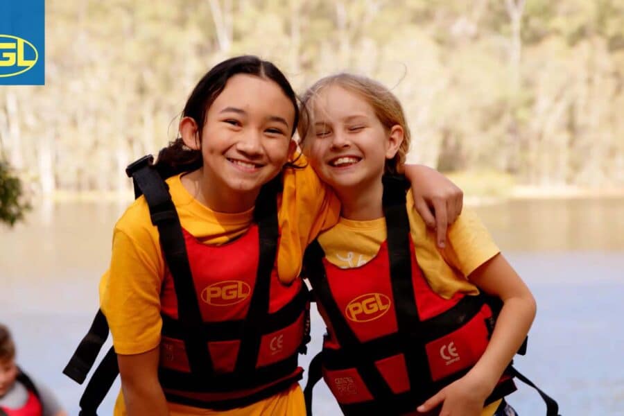 Two smiling girls wearing yellow life jackets and backpacks by a river, with a PGL holiday camps logo on their life jackets.