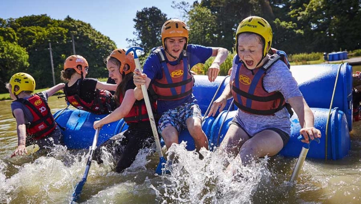 A group of young people wearing helmets and life jackets enthusiastically paddle a raft through splashing water on a sunny day.