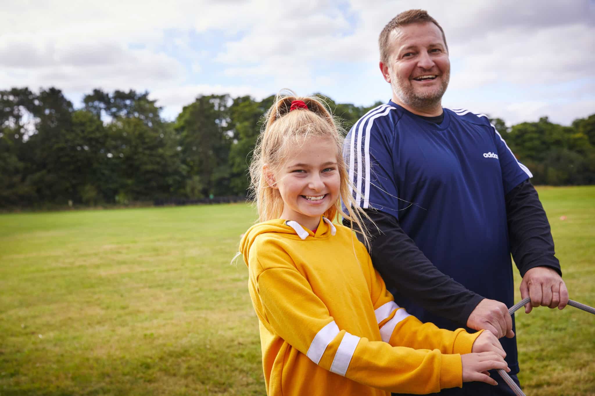 A smiling girl in a yellow hoodie and a man in a blue athletic shirt stand outdoors on a grassy field, both holding long handles, enjoying the activities typically found at group camps under a partly cloudy sky.