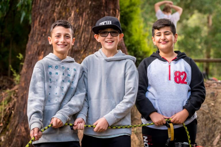 Three boys stand outdoors, smiling and holding a rope, with a large tree trunk and greenery in the background. One boy wears a cap and glasses. Their joyful expressions capture the spirit of group camps perfectly.