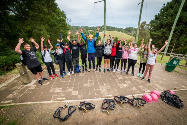 A group of people outdoors raises their hands in excitement, standing on a paved area with trees and hills in the background. Safety harnesses and helmets are on the ground in front of them.