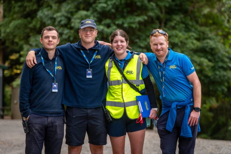 Four people in branded uniforms and lanyards stand outdoors, with two wearing hats and one in a yellow safety vest holding a clipboard. Trees are visible in the background.