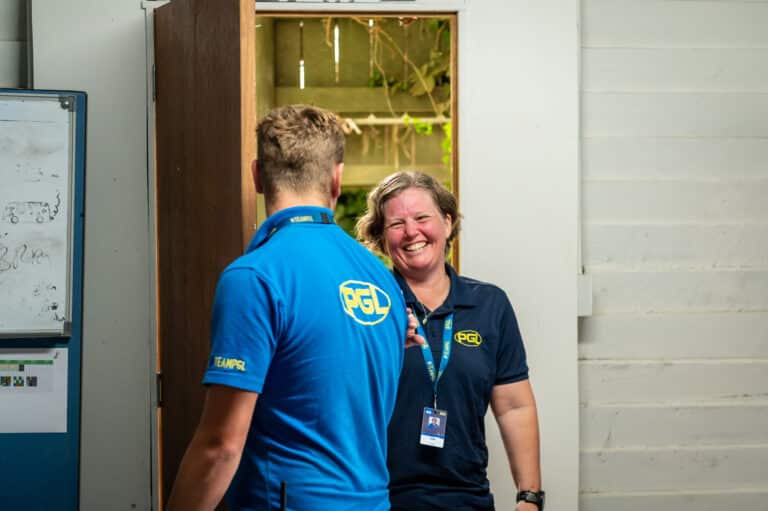 Two people wearing blue PGL shirts chat and smile in a room with an open door. One person has their back to the camera.